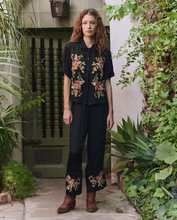 A woman stands outdoors among green plants, wearing THE GREAT. PF25 The Tavern Button Up—black with bouquet floral embroidery—and pants, paired with brown boots.