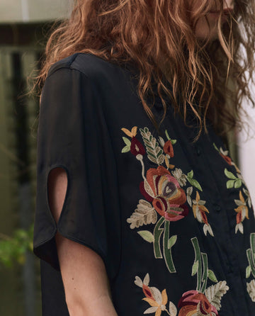 A person with wavy brown hair wears THE GREAT. PF25 The Tavern Button Up in black, featuring colorful bouquet floral embroidery and short sleeves with a slit.