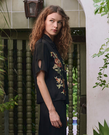 A woman with curly hair stands outdoors by a green gate, wearing THE GREAT. PF25's The Tavern Button Up in black with bouquet floral embroidery, surrounded by plants.