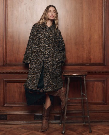 A woman in THE GREAT. F26 COLLECTORS' The Trench Coat. -- Dark Leopard pairs it with brown boots, standing by a wooden stool against a paneled wall, embodying the bold elegance of women's outerwear.