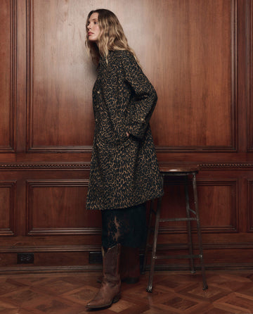 A woman wearing The Trench Coat in Dark Leopard by THE GREAT. F26 COLLECTORS, paired with a black skirt and brown cowboy boots, stands beside a wooden stool against a paneled wood wall.