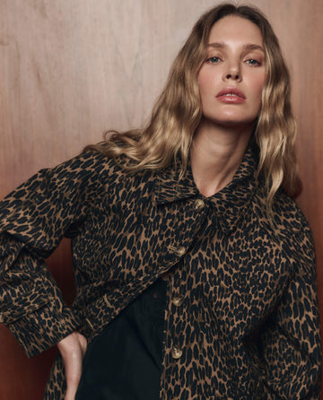 A person with long wavy hair poses in front of a wooden background, wearing THE GREAT. F26 COLLECTORS' The Trench Coat in Dark Leopard—a bold statement in women's outerwear.