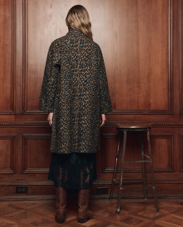A person with long hair stands facing a wooden wall, wearing THE GREAT. F26 COLLECTORS The Trench Coat in Dark Leopard, paired with a black lace skirt and brown boots; a wooden stool is beside them.