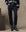 Wearing THE GREAT. H25 COLLECTION’s The Satin Wayne pants in black, paired with a black patterned sweater, a statement belt, and black cowboy boots, a person stands by a wooden box against a neutral backdrop.