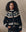 A woman with curly hair models THE GREAT. H25 COLLECTION’s The Snowflake Ice Rink Cardigan in black over a white collared shirt, standing against a neutral backdrop.