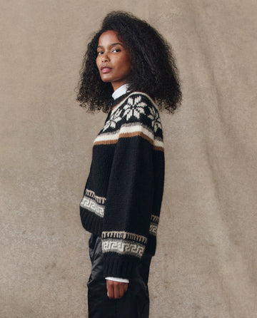 A woman with curly hair stands in profile wearing THE GREAT. H25 COLLECTION's Snowflake Ice Rink Cardigan in black, featuring white and beige geometric patterns, paired with black pants against a neutral background.