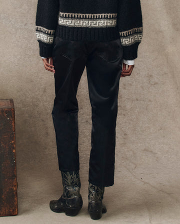A person stands facing away, wearing The Satin Wayne in black by THE GREAT. H25 COLLECTION—a black and white patterned sweater, black pants, and foliage-detailed boots—beside a wooden box. The satin’s subtle sheen shines against a neutral backdrop.