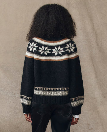 A person with curly hair is seen from the back wearing THE GREAT. H25 COLLECTION’s The Snowflake Ice Rink Cardigan in black, featuring white snowflake and geometric designs, photographed against a neutral background.