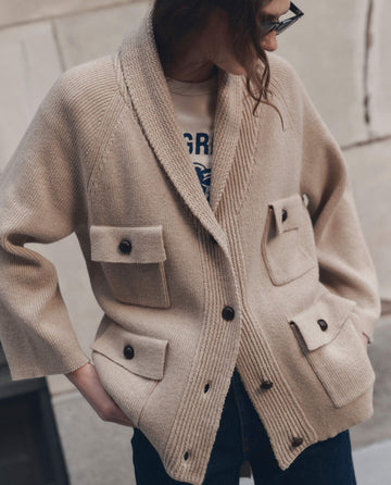 A person wearing THE GREAT. F26 D1 The Commander Riding Cardigan in Cream, styled with a printed t-shirt and dark pants, stands against a beige wall.