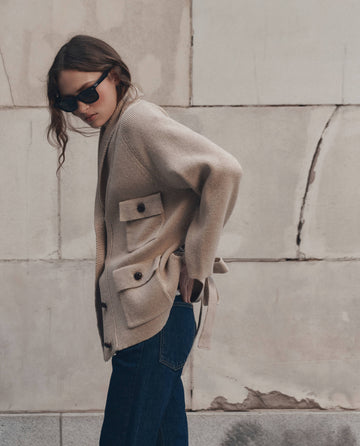 Wearing sunglasses, a woman stands against a light-colored, textured wall in THE GREAT.’s The Commander Riding Cardigan in Cream, styled with its large pockets and paired with dark jeans.