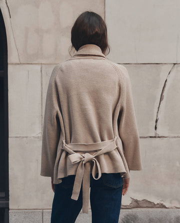 A person with their back to the camera stands facing a stone wall, wearing THE GREAT. F26 D1's The Commander Riding Cardigan in cream and dark jeans.