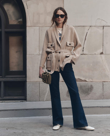 A woman stands outdoors wearing sunglasses, a cream The Commander Riding Cardigan by THE GREAT. F26 D1 belted over a white shirt, paired with dark blue jeans and white shoes, and holding a small striped bag in her left hand.