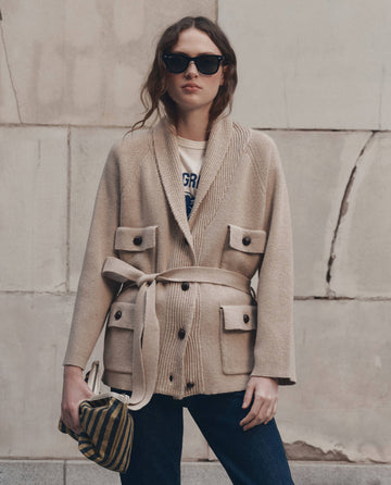 A person wearing sunglasses, THE GREAT. F26 D1 The Commander Riding Cardigan in Cream with large pockets, a white printed shirt, and blue jeans stands against a stone wall, holding a striped bag.
