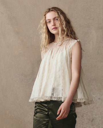 A person with long wavy blonde hair wears THE GREAT. H25 COLLECTION’s The Keepsake Lace Shell Top in cream and dark green pants, standing against a neutral textured background.