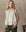 A woman with wavy blonde hair wears THE GREAT. H25 COLLECTION’s The Keepsake Lace Shell Top in Cream and olive green pants, standing by a wooden table against a neutral backdrop.