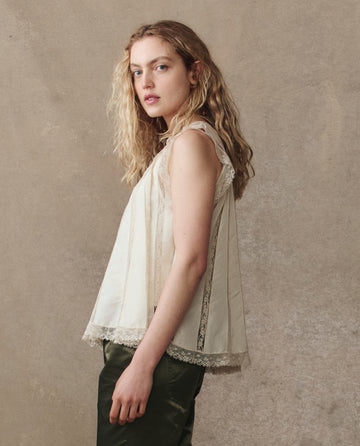 A woman with long wavy blonde hair wears THE GREAT. H25 COLLECTION's Keepsake Lace Shell Top in cream, paired with dark green pants, standing before a beige textured background.
