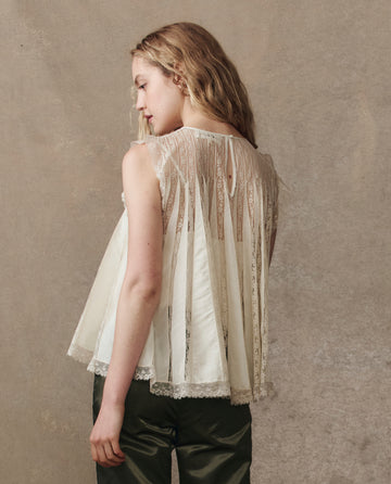 A person stands facing away, wearing THE GREAT. H25 COLLECTION’s The Keepsake Lace Shell Top in cream with vertical lace panels and dark green pants, set against a neutral background.
