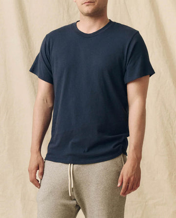 A man stands against a beige fabric backdrop wearing THE GREAT. FALL 23 MEN's The Men's Boxy Crew in True Navy—a true wardrobe staple.