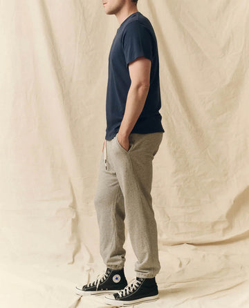 A person stands in profile, hands in pockets, wearing THE GREAT. FALL 23 MEN The Men's Boxy Crew. -- True Navy, gray sweatpants, and black high-top sneakers against a beige fabric backdrop.