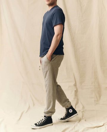 A person stands sideways with hands in pockets, wearing THE GREAT. FALL 23 MEN The Men's Boxy Crew in True Navy—styled with gray jogger pants and black high-top sneakers—against a beige fabric backdrop.