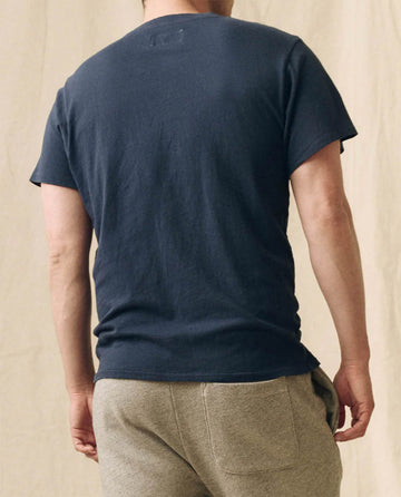 A person stands with their back to the camera, wearing THE GREAT. FALL 23 MEN The Men's Boxy Crew in True Navy and light gray sweatpants against a neutral background.