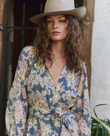 A woman in THE GREAT. PF25’s Indigo Floral Pampas Dress stands gracefully before a wrought iron gate and stone wall.