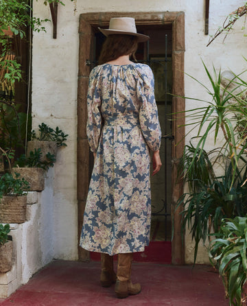 Wearing THE GREAT. PF25 The Pampas Dress in Indigo Floral with a beige hat and brown boots, a person stands facing a rustic door framed by pampas and greenery.
