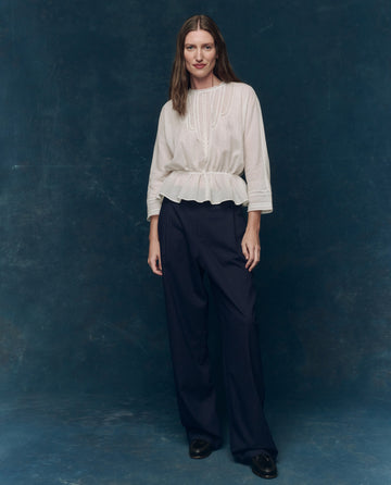 Against a dark blue backdrop, a person wears THE GREAT. F25 COLLECTORS' The Victorian Top in white, styled with dark wide-leg trousers and black shoes.