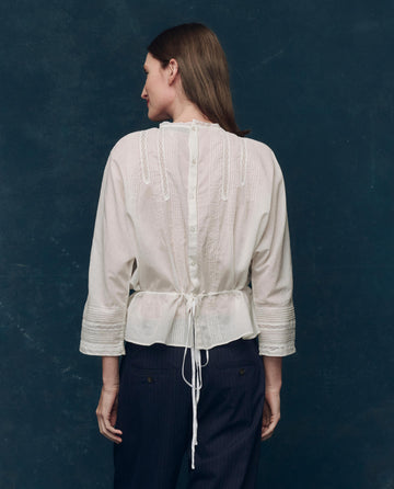 A person stands facing away, wearing THE GREAT. F25 COLLECTORS The Victorian Top in white— a vintage-inspired cotton voile blouse with button and tie details—paired with dark pants against a textured backdrop.