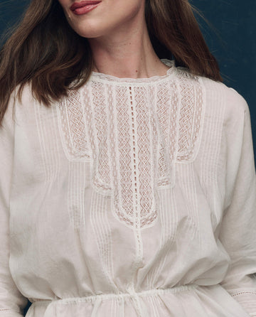 A woman stands against a dark background wearing The Victorian Top in white by THE GREAT. F25 COLLECTORS—a vintage-inspired, long-sleeved cotton voile blouse with intricate lace detailing that adds elegance to her look.