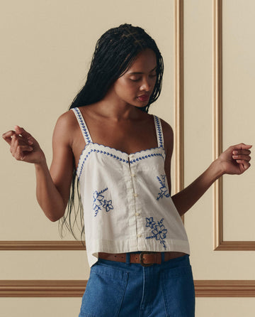 A woman models THE GREAT. SP26 D2 The Weekend Tank W/ Hanky Embroidery in cream and blue jeans, standing against a beige wall—an ideal relaxed weekend look.