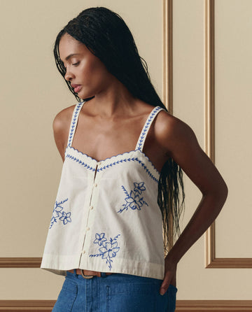 A woman wearing THE GREAT. SP26 D2 The Weekend Tank W/ Hanky Embroidery in cream and blue jeans stands before a beige paneled wall, looking down and to the side.