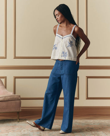 A woman stands indoors in THE GREAT.’s The Weekend Tank W/ Hanky Embroidery in cream, paired with wide-leg blue jeans and beige flats, beside an upholstered chair against a beige paneled wall.