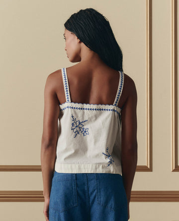 A woman stands with her back to the camera, wearing THE GREAT.'s The Weekend Tank W/ Hanky Embroidery in cream and blue pants, set against a beige wall with decorative molding.