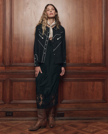 A woman stands indoors against wood paneling, wearing THE GREAT. F26 COLLECTORS' The Western Shirt in black, styled in classic western fashion with a black lace-trimmed dress, boots, and a light scarf tied around her neck.