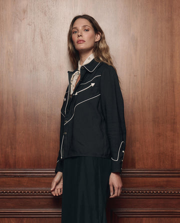 A person with long hair, dressed in THE GREAT. F26 COLLECTORS' The Western Shirt in Black—a black jacket with white piping that evokes a classic western vibe—and a light scarf, stands before a wooden paneled wall.