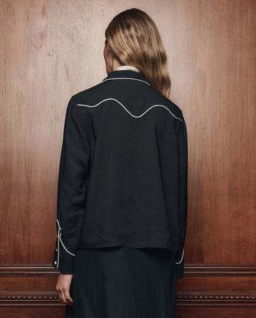 A person with long, wavy hair stands facing a wooden wall, wearing THE GREAT. F26 COLLECTORS The Western Shirt in black with white piping details and matching black pants.