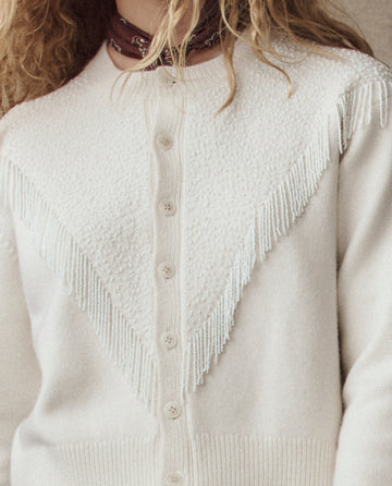 A person wearing THE GREAT. H25 COLLECTION's The Beaded Fringe Cardigan in cream, featuring textured and fringed detailing on the chest, paired with a bandana around their neck.