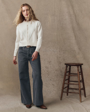 A person with long blonde hair wears THE GREAT. H25 COLLECTION's The Beaded Fringe Cardigan in Cream with wide-leg jeans, standing by a wooden stool against a neutral backdrop.