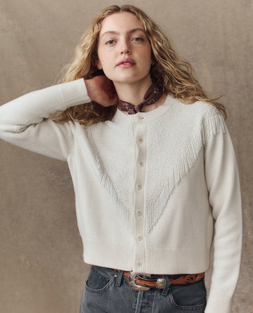 Wearing THE GREAT. H25 COLLECTION's The Beaded Fringe Cardigan in Cream, a person with long curly hair, a brown bandana, and dark belted jeans stands against a neutral background.