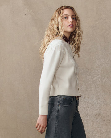 A woman with long curly blonde hair stands sideways against a textured beige background, wearing THE GREAT. H25 COLLECTION's The Beaded Fringe Cardigan in cream with dark jeans.