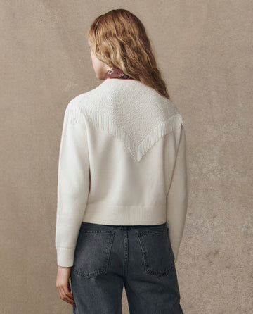 A person with long, wavy hair wears THE GREAT. H25 COLLECTION's The Beaded Fringe Cardigan in cream, featuring beaded fringe and textured back detailing, paired with high-waisted grey jeans against a beige background.