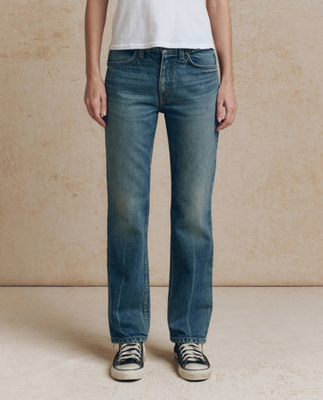 A person wearing THE GREAT. F26 D1 The Clark Mid-Rise Straight Jean in Fairfield Wash With Bleach Crease, paired with a white t-shirt and black sneakers, stands against a beige textured wall.