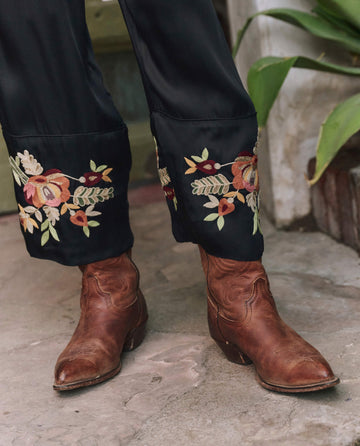 A person wears THE GREAT. PF25’s The Cuffed Pant in black with Bouquet Floral Embroidery, paired with brown leather cowboy boots, standing on a concrete floor by green plants.