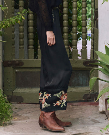 A person wearing THE GREAT. PF25 The Cuffed Pant in black with bouquet floral embroidery stands on a stone porch near green plants and a wooden door.