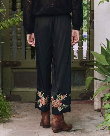 A person stands outdoors facing away, wearing THE GREAT. PF25 The Cuffed Pant in black with bouquet floral embroidery at the cuffs and brown boots; plants and a green wooden gate in the background.