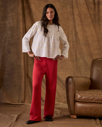 A person wearing a white long-sleeved blouse with lace detailing and red The Fleece Lap Sweatpant. -- Campfire, standing next to a leather armchair.