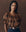 A woman with long braids wears THE GREAT. F25 D1's The Moorland Top in Geo Bandana Print, paired with navy pinstripe pants and a decorative belt, standing against a plain brown background.