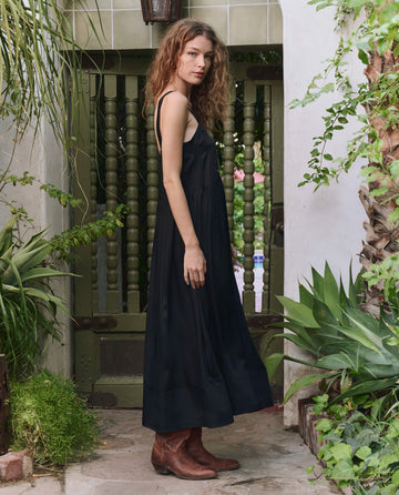 A woman with curly hair stands outdoors by a green wooden gate, wearing THE GREAT. PF25 The Day Dress in black with bouquet floral embroidery and brown boots, surrounded by lush plants.