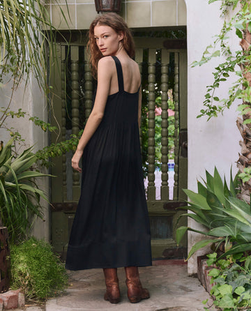 A woman with long hair wears THE GREAT. PF25's The Day Dress in black with bouquet floral embroidery, styled with brown boots, as she stands outdoors near a green gate surrounded by plants, looking over her shoulder.